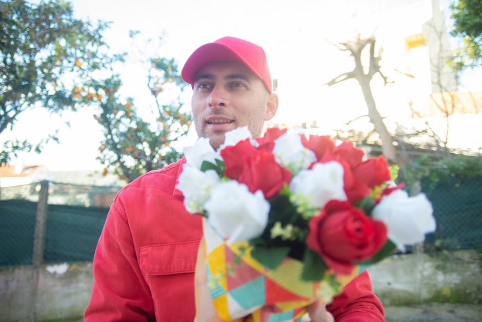 A delivery person dressed in a red uniform with a matching red cap, holding a vibrant bouquet of fresh flowers. The bouquet includes red and white roses, with green foliage, wrapped in colorful patter
