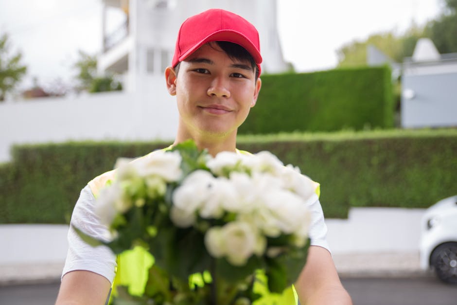 A delivery person wearing a red cap and a neon yellow vest is standing outdoors, holding a bouquet of fresh white flowers with green leaves. The flowers appear to be roses or peonies with vibrant, ful