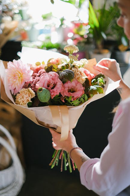 A person holding a fresh floral bouquet featuring large pink peonies, creamy carnations, and white hydrangeas, accented with dark green leaves, wrapped in white tissue paper and tied with a cream-colo