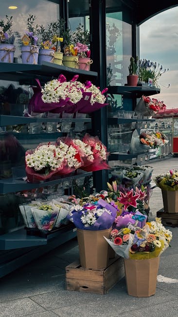 A display of fresh floral bouquets and arrangements on metal shelving inside a flower shop with large glass windows. The bouquets feature a variety of flowers including white chrysanthemums, pink carn