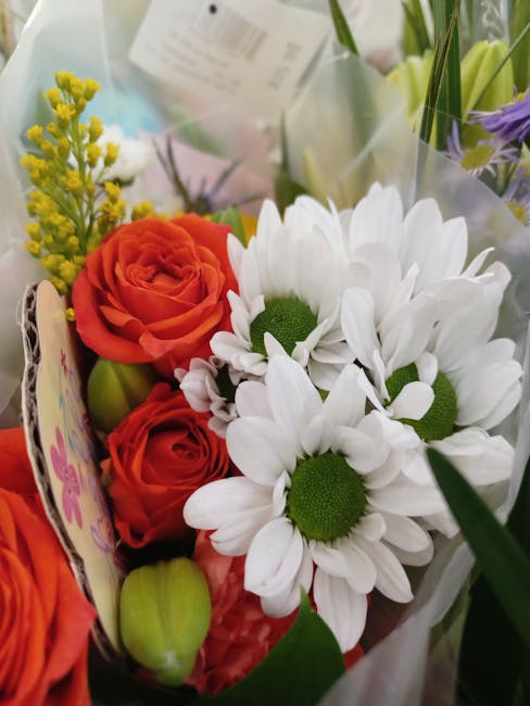 A vibrant floral bouquet featuring large white daisies with green centers, complemented by bright orange roses with tightly layered petals, and small yellow mimosa blossoms on delicate green stems. Th