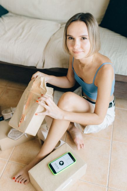 A young woman with shoulder-length blonde hair and fair skin is sitting on a tiled floor next to a beige sofa with a black pillow. She is wearing a light blue spaghetti strap top and white shorts. In