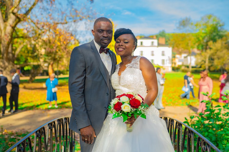 A bride and groom stand together outdoors on a sunny day, with the bride holding a floral bouquet featuring large red and white roses, accented by green leaves and delicate filler flowers. The bride w